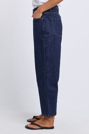 A person stands in profile against a plain light background wearing Foxwood Ambrose Barrel Jean, a dark blue high-waisted stretch denim, with one hand in their pocket and black sandals.