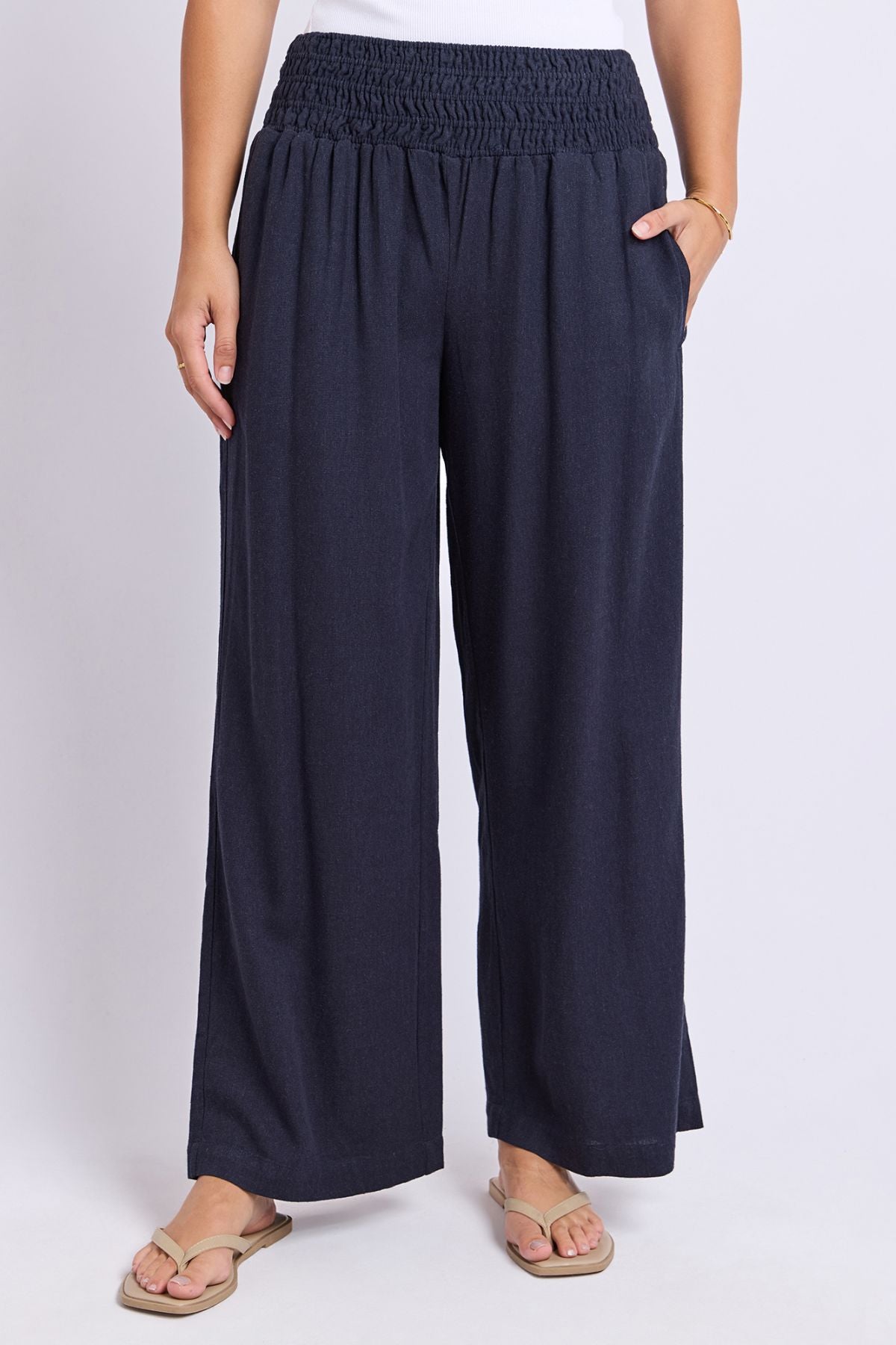 Wearing the Foxwood Amie Pant in navy blue with a smocked waistband, paired with a white top and beige flip-flops, this person stands against a plain light background with hands in their pockets.
