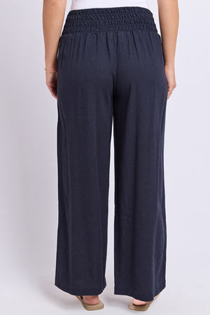 A person is shown from the lower back down, standing with their back to the camera against a plain light background, wearing the Foxwood Amie Pant—loose-fitting navy blue pants featuring a smocked elastic waistband.