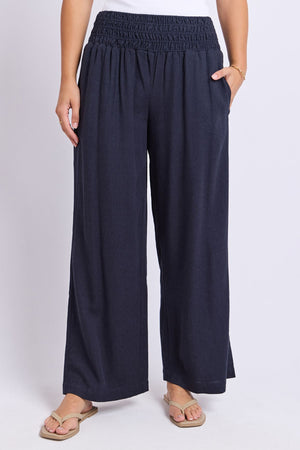 Wearing the Foxwood Amie Pant in navy blue with a smocked waistband, paired with a white top and beige flip-flops, this person stands against a plain light background with hands in their pockets.