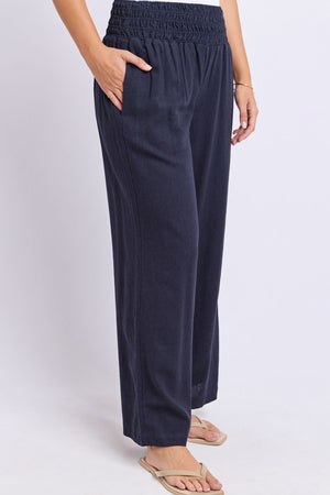 Wearing the Foxwood Amie Pant in a loose-fitting, dark navy wide-leg style with an elastic waistband, paired with tan flip-flops and a thin bracelet, this person stands with one hand in their pocket against a plain light background.