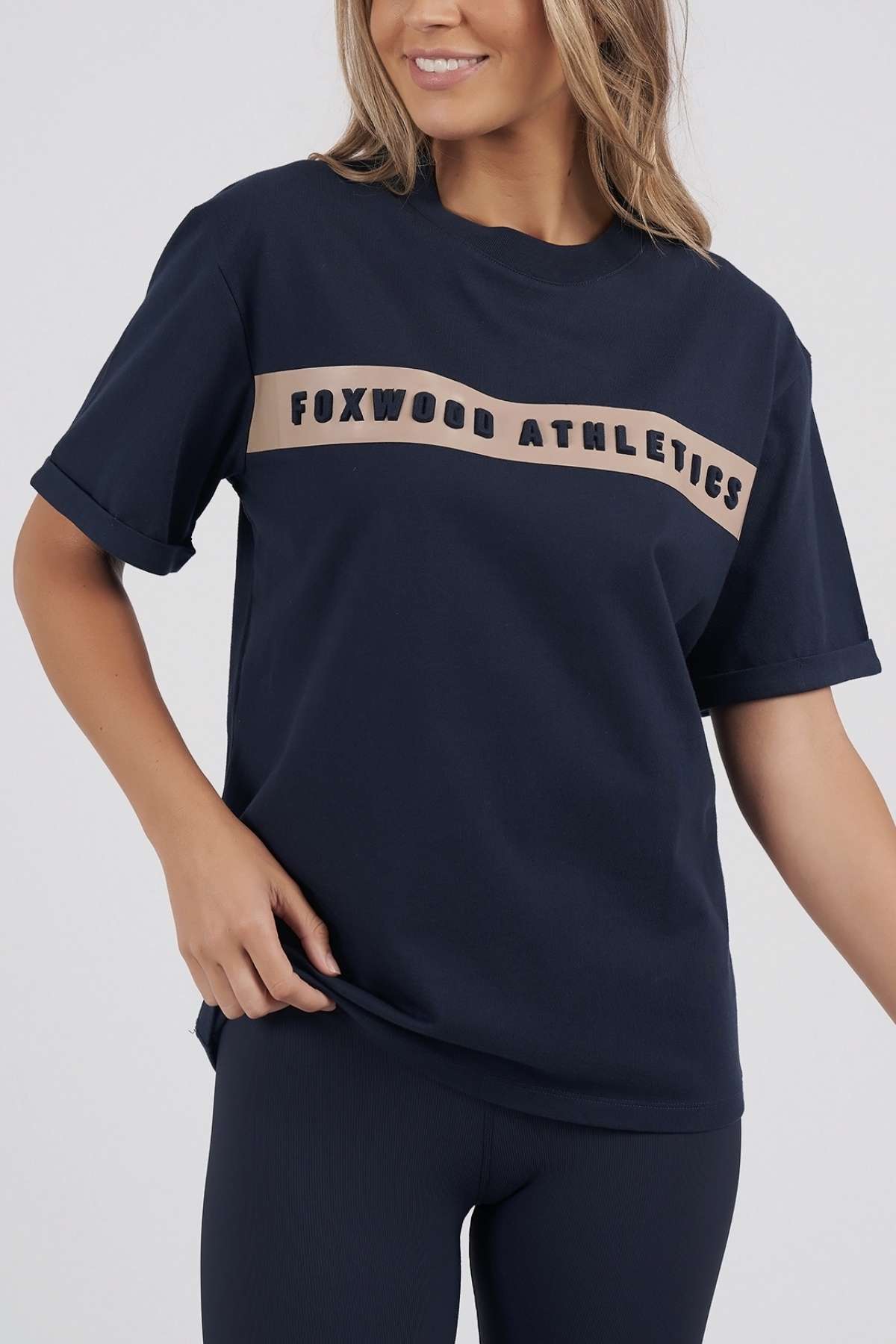 Against a simple backdrop, an individual with long, wavy hair wears a navy Foxwood Athletics Tee with dark leggings. The tee showcases a beige stripe across the chest embossed with "Foxwood Athletics," completing the casual yet stylish ensemble.