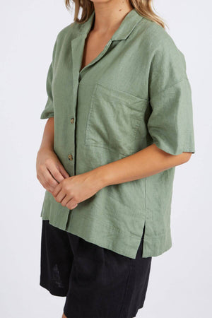 A person wears a loose-fitting Foxwood Aubrey shirt in light green linen, featuring a button-through placket and short sleeves, paired with black shorts. Their light-colored hair contrasts with the plain background.