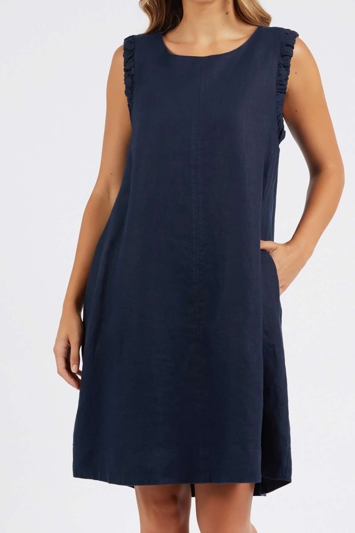 A woman models the Foxwood Bastia Dress, a navy blue, sleeveless A-line dress with ruffled shoulders and pockets, standing against a plain light background.