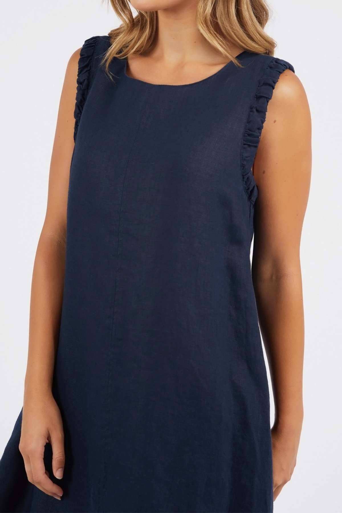 A woman stands against a plain white background wearing the Foxwood Bastia Dress, a sleeveless navy blue A-line style with ruffled shoulder detailing. Her face is partially out of frame.