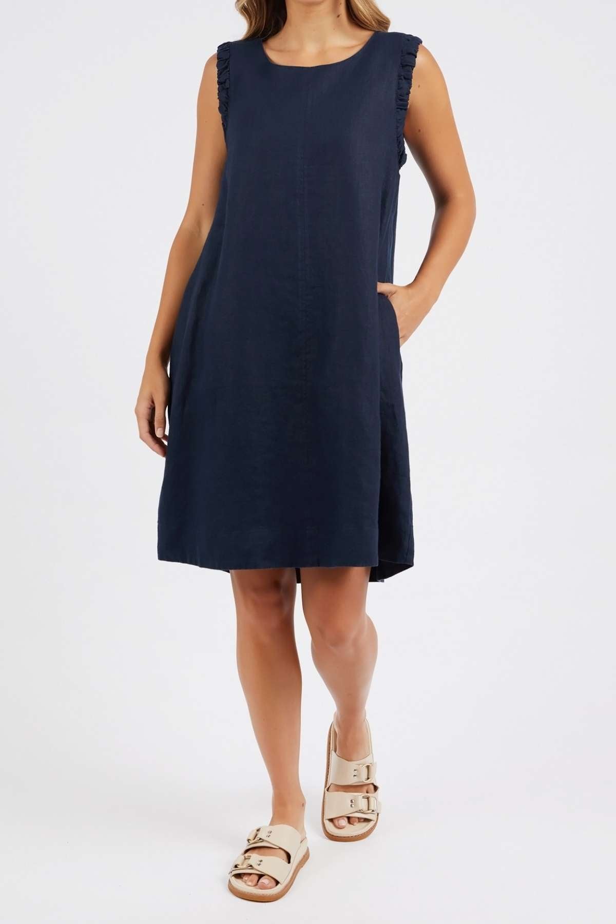 A woman stands with one hand in her pocket against a plain white background, wearing the Foxwood Bastia Dress—an A-line, sleeveless knee-length navy linen dress with ruffled shoulder details—and beige sandals.