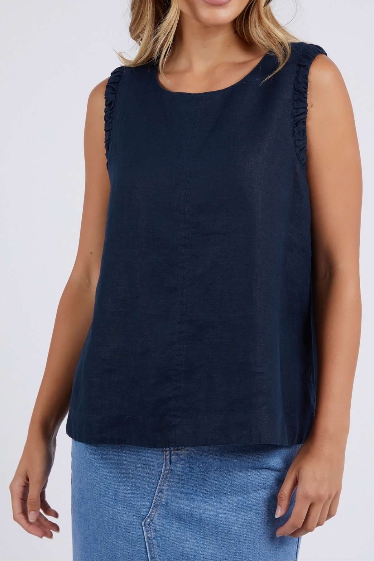 A woman wears the Foxwood Bastia Top—navy blue, 100% linen with ruffled shoulders—paired with a light blue denim skirt, standing against a plain background.