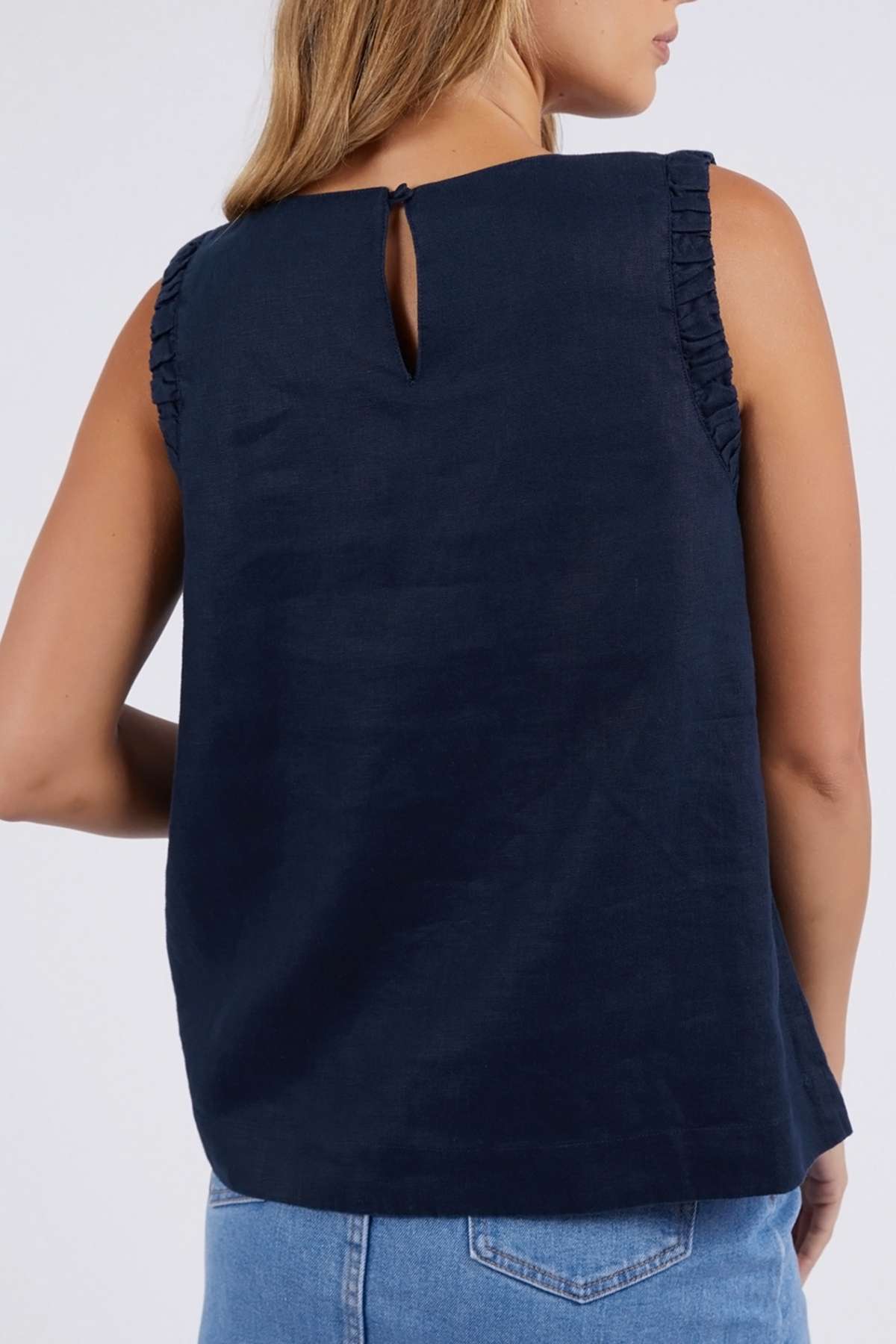 A woman is seen from behind wearing the Foxwood Bastia Top—a relaxed-fit, navy sleeveless top with ruffled armholes and a keyhole neckline—paired with light blue jeans.
