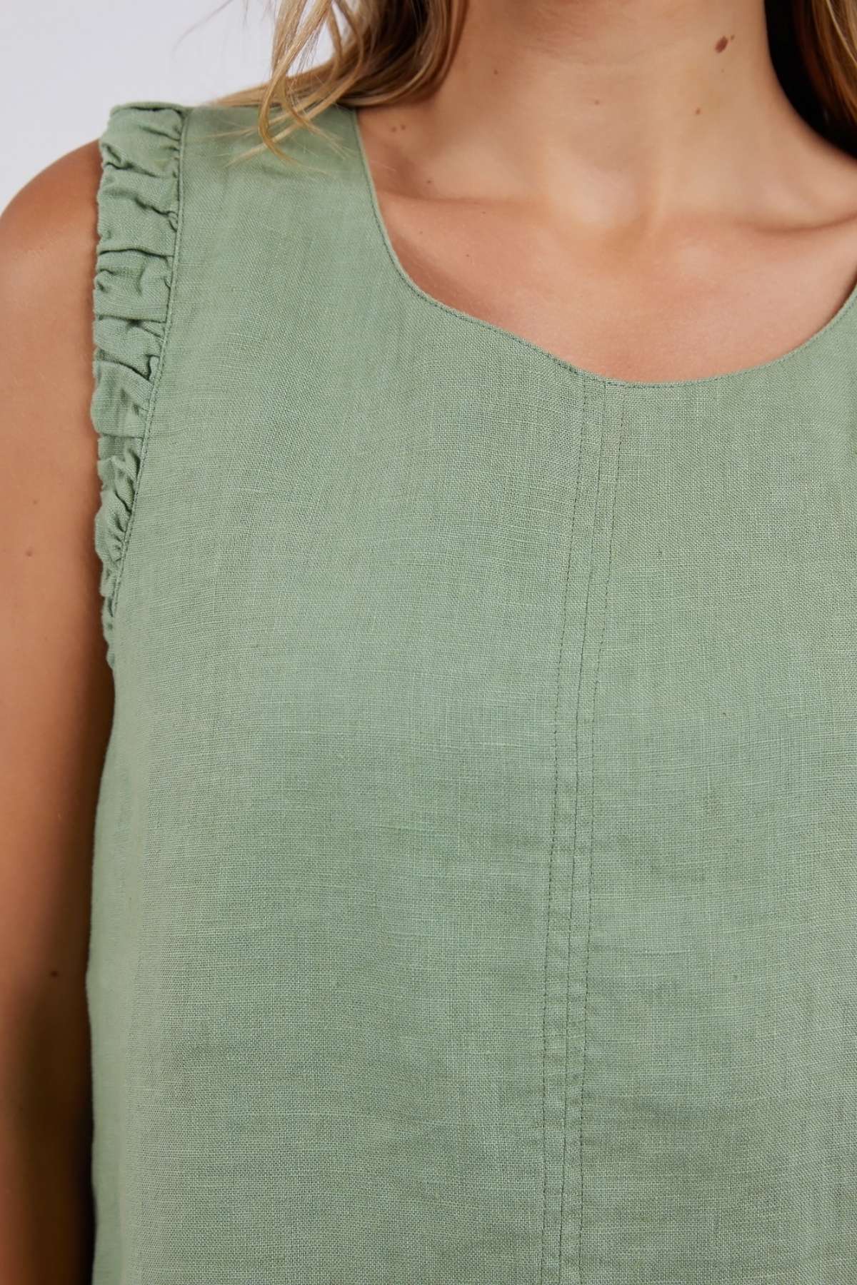 A person wears the Foxwood Bastia Top, a sleeveless, light green linen garment with gathered shoulder ruffles. Visible stitching adorns the front, and their shoulder-length light brown hair cascades over one shoulder.