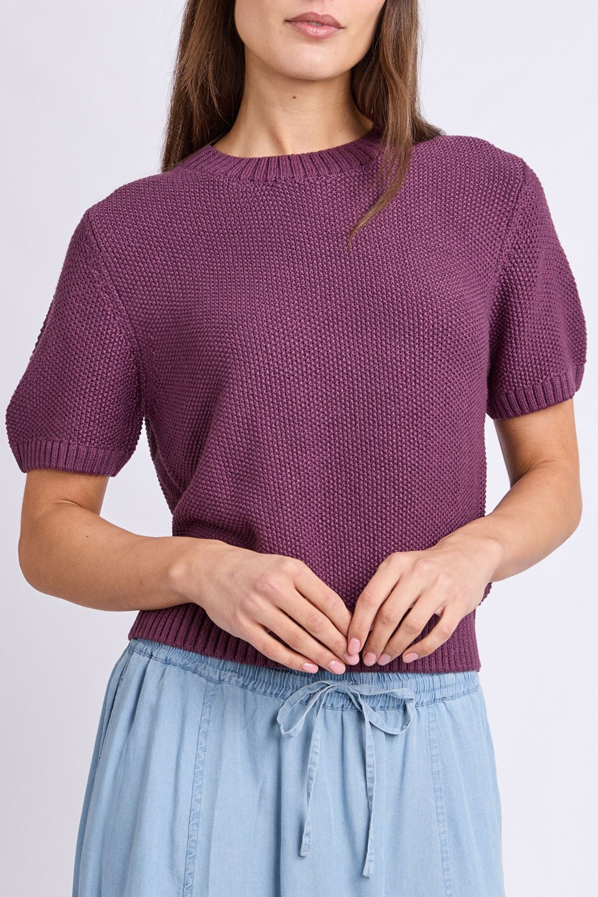 A woman wearing the Foxwood Blair Short Sleeve Knit in textured purple and light blue drawstring pants stands with her hands clasped at her waist against a plain background.
