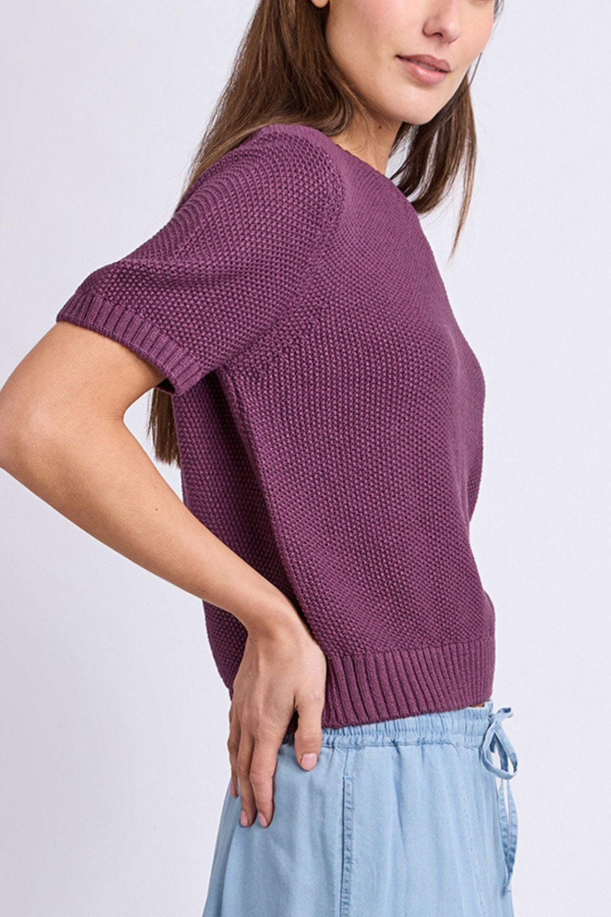 A woman wearing the Foxwood Blair Short Sleeve Knit in a textured purple finish with light blue drawstring pants, standing sideways with one hand on her hip against a plain background.