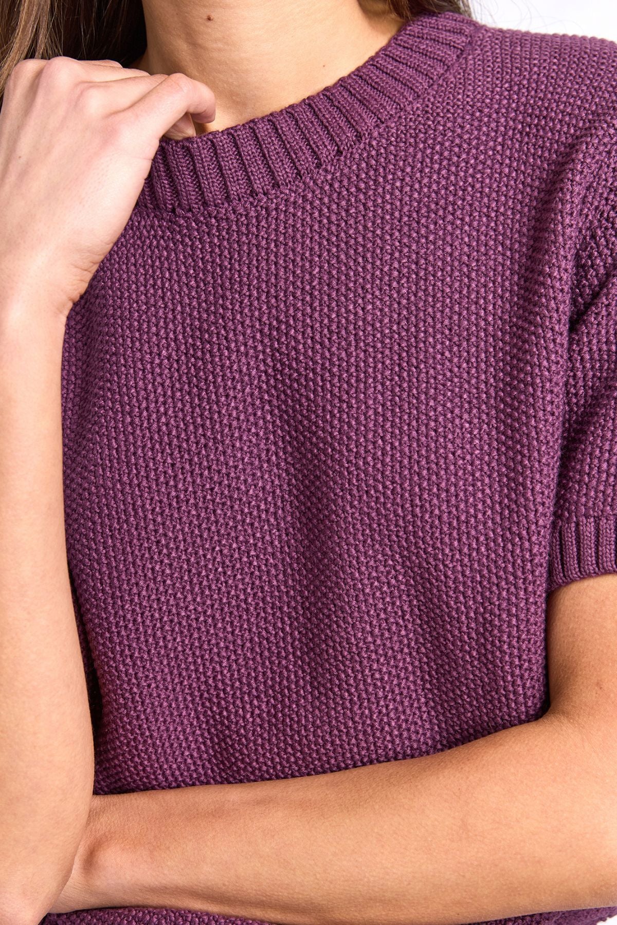 A person wearing the Foxwood Blair Short Sleeve Knit in purple, featuring a textured design, rests one arm across their body while touching their chin with the other hand.