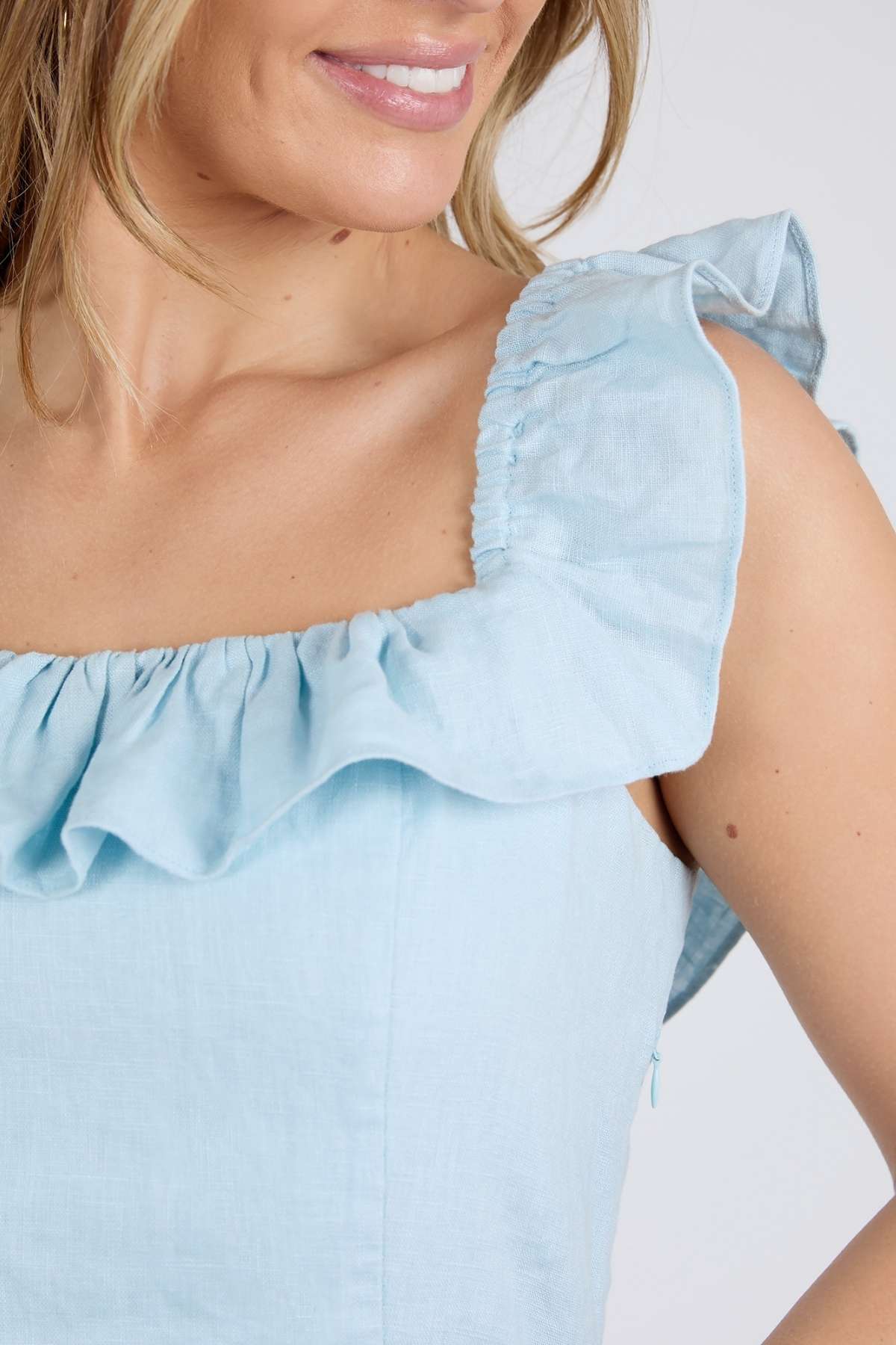 A woman smiles softly in the Foxwood Bonifacio Dress, showcasing its light blue linen fabric and ruffled sleeveless neckline, with focus on the upper part of the dress and her shoulder.