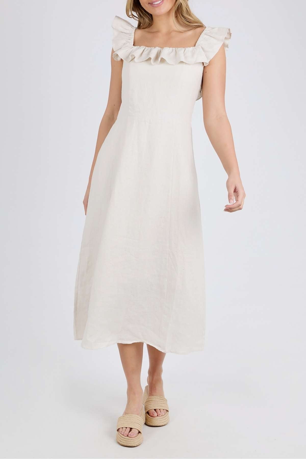 A woman in the Foxwood Bonifacio Dress—light beige linen with a ruffled square neckline—stands before a white background, her arm slightly bent and hair worn loose.