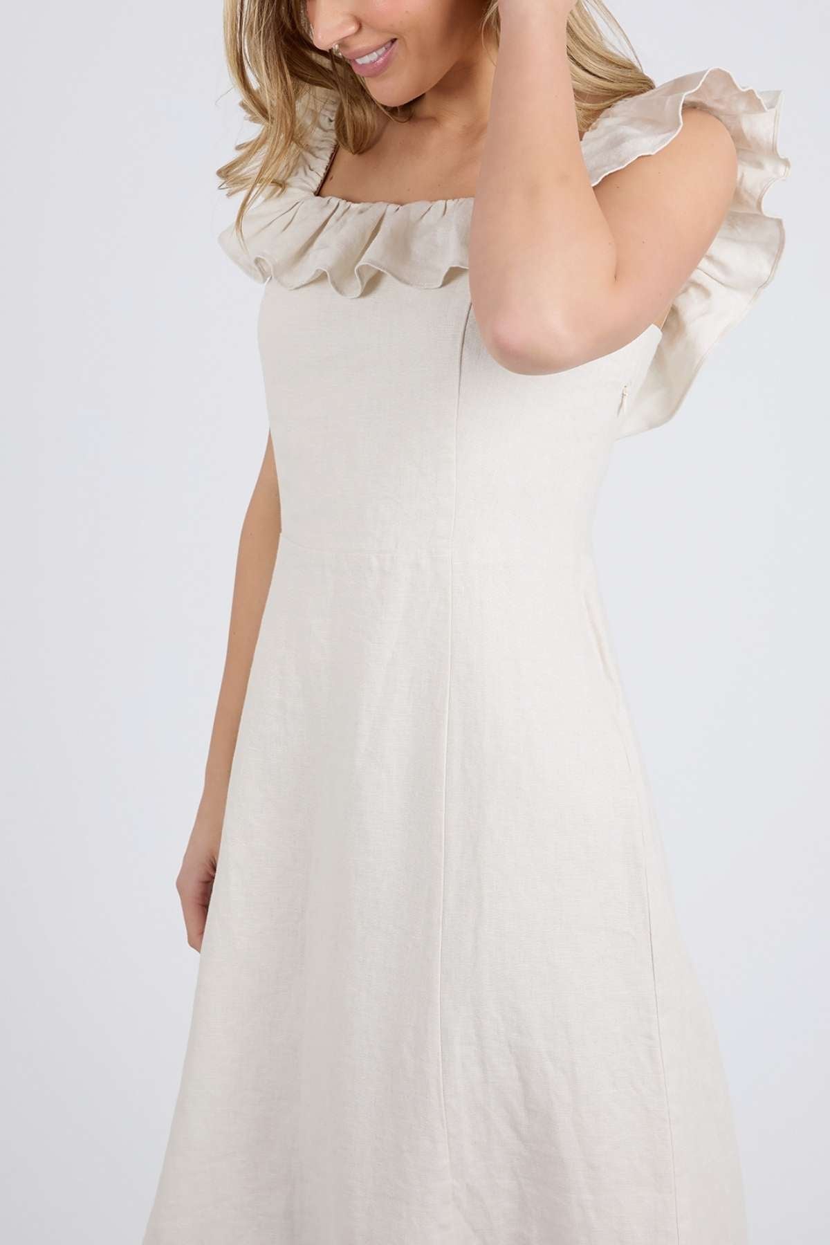 A woman poses against a plain backdrop in the off-shoulder Foxwood Bonifacio Dress, featuring ruffled sleeves. Her light brown hair partially obscures her face as she elegantly touches her hair, embodying effortless luxury.