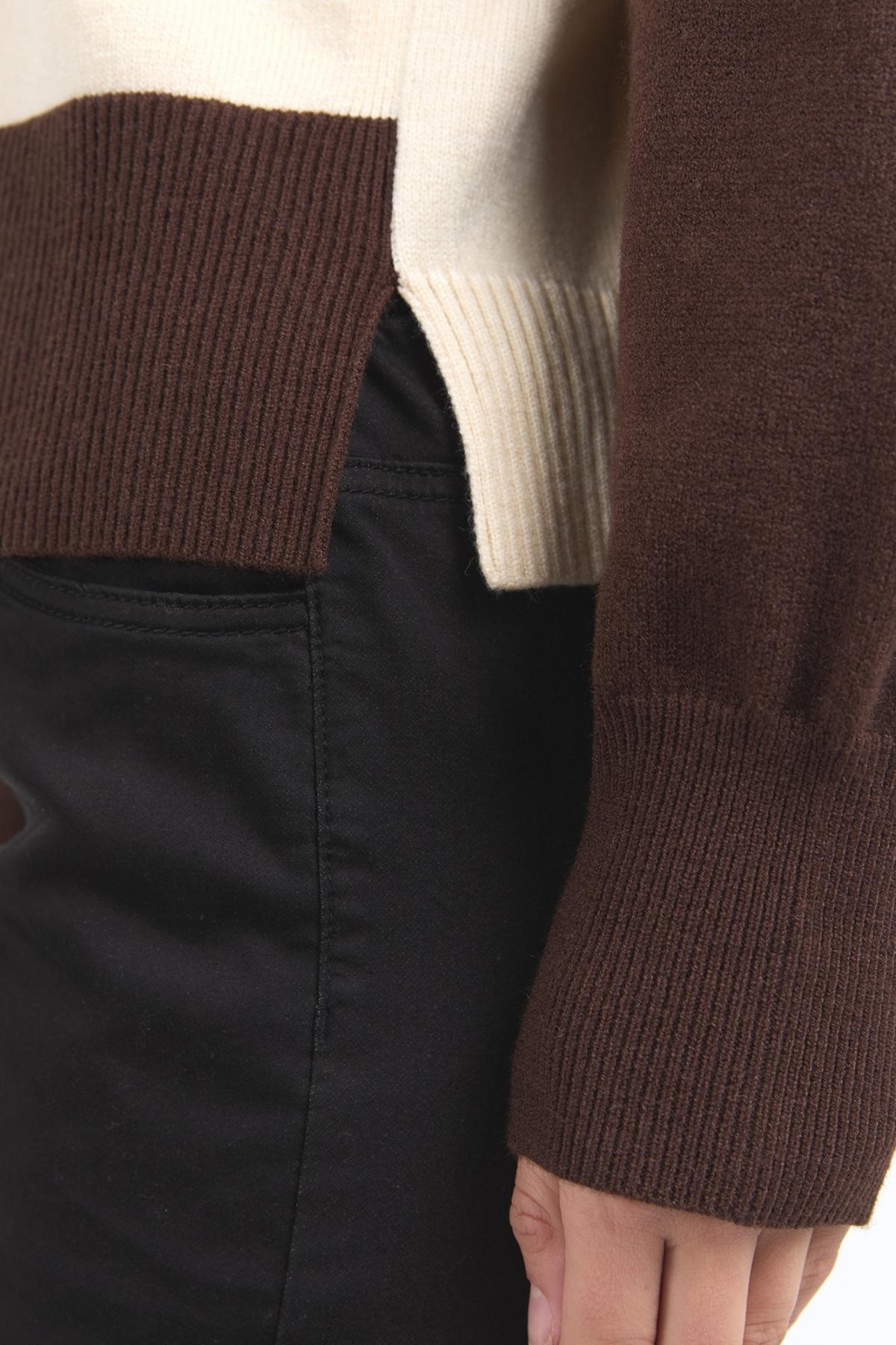 A close-up shows a person in black pants and the Foxwood Canterbury Knit, featuring cream and brown sections, ribbed cuffs and hem, a side slit at the bottom, and their hand relaxed at their side.