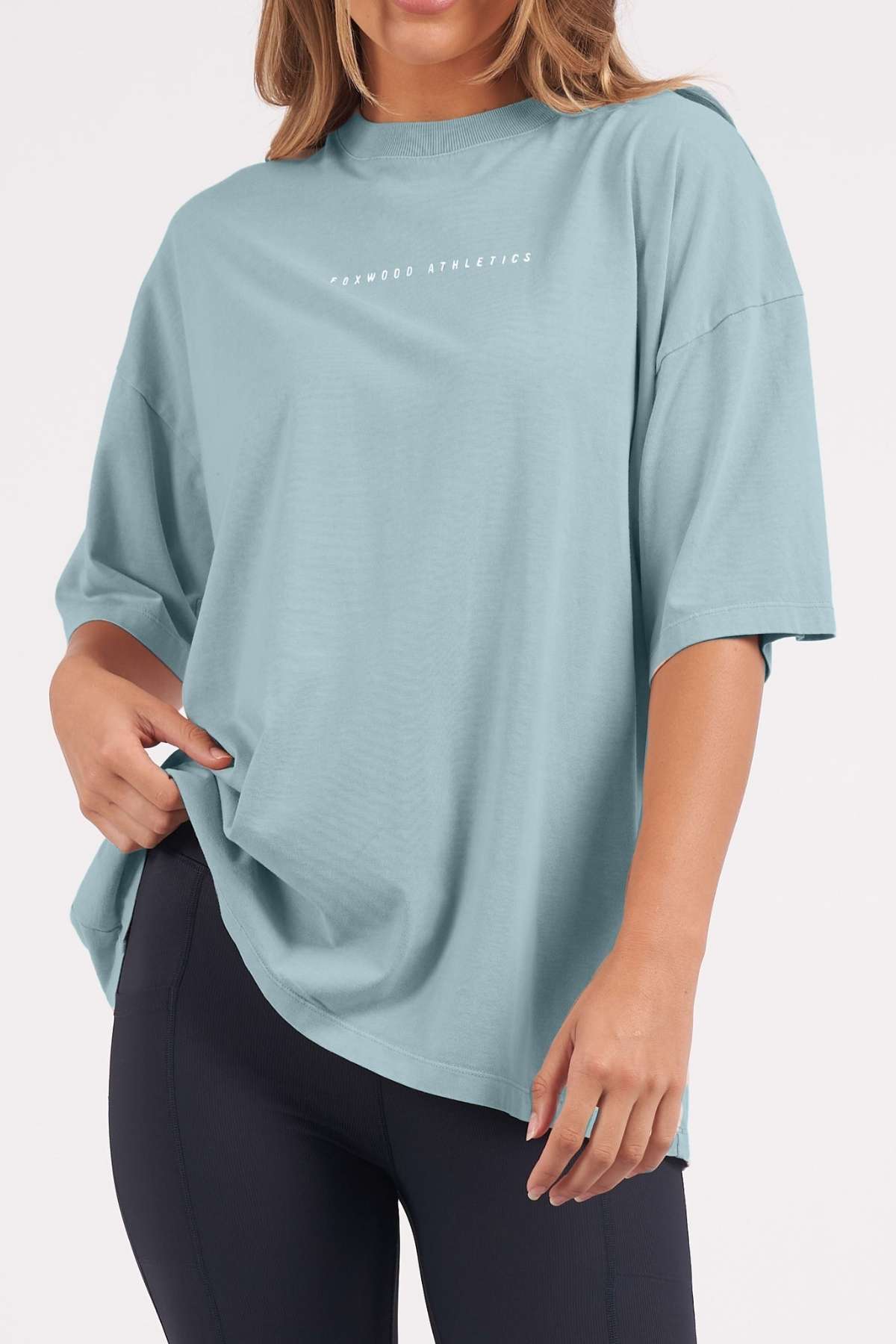 A woman wears the Foxwood Captain Tee, a casual light blue cotton top with "Foxwood Athletics" on the chest, styled oversized with black leggings and one hand holding the hem for a relaxed look.