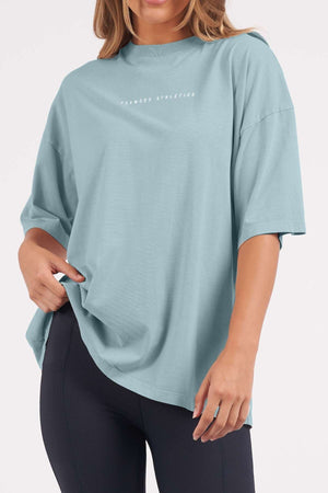A woman wears the Foxwood Captain Tee, a casual light blue cotton top with "Foxwood Athletics" on the chest, styled oversized with black leggings and one hand holding the hem for a relaxed look.