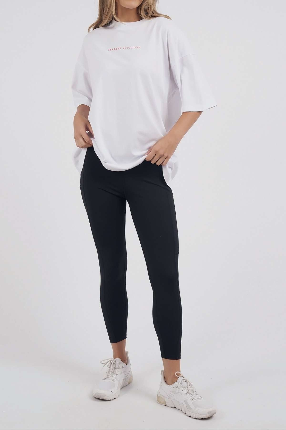 A person stands against a plain white background wearing the oversized Foxwood Captain Tee in white, paired with black leggings and white sneakers. The casual look is perfect for everyday wear. The person's face is not visible in the image.