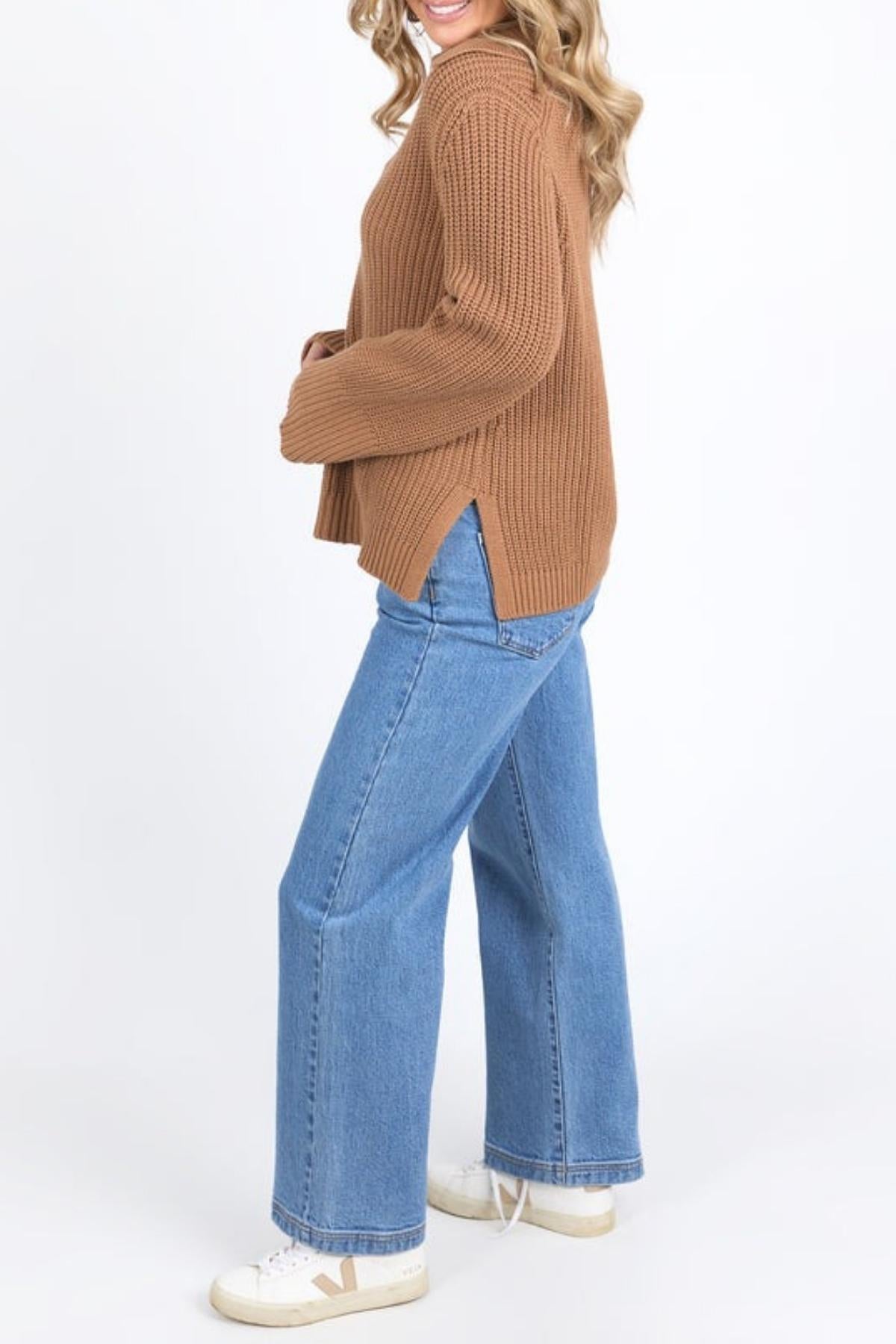 Wearing the Foxwood Carlton Knit brown sweater, blue wide-leg jeans, and white sneakers, a person stands sideways against a plain white background, effortlessly highlighting comfort and style.
