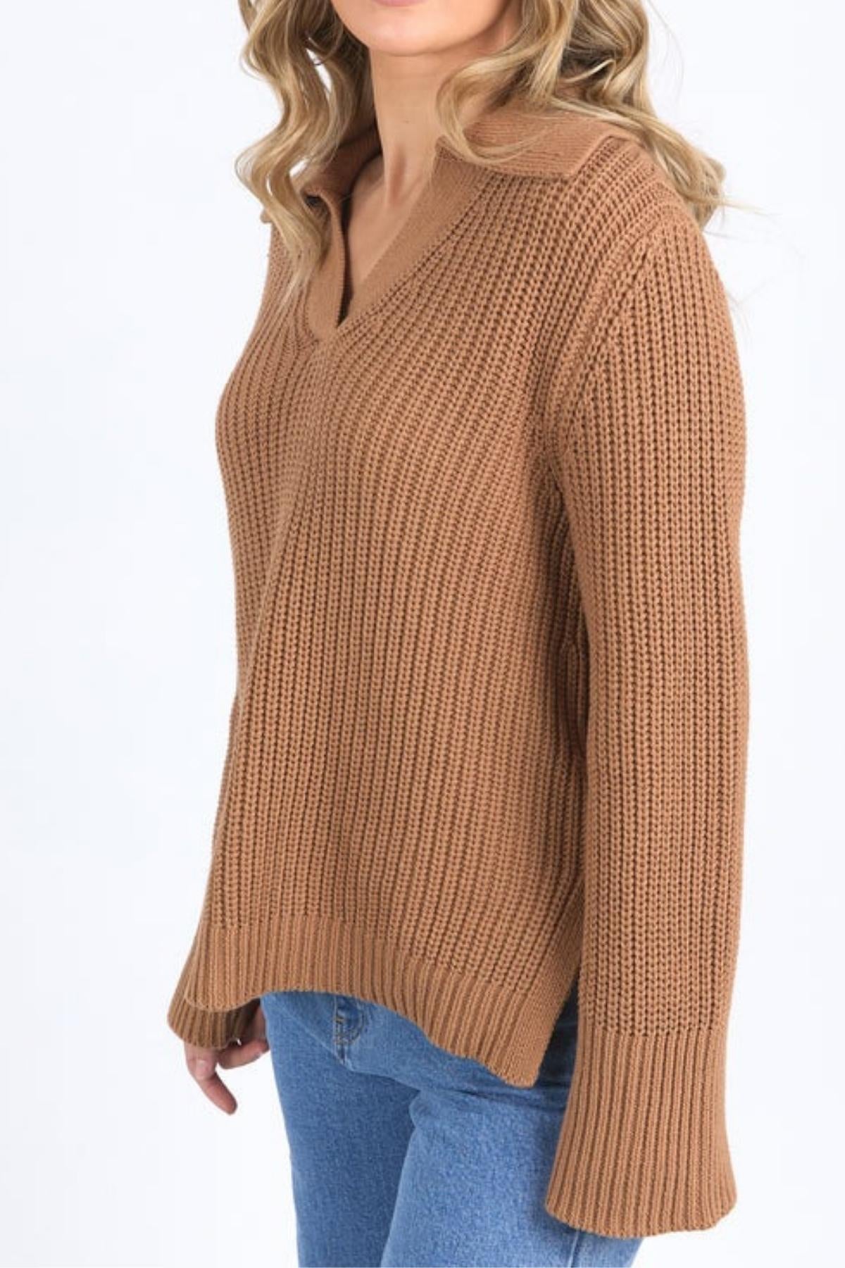 A person with wavy blonde hair wears the Foxwood Carlton Knit—a brown, relaxed-fit knit sweater with a collar—styled with blue jeans, standing against a white background. The partially visible face adds to the look’s effortless comfort and style.