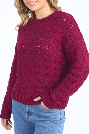 A woman smiles slightly, standing with one hand in her pocket, wearing blue jeans and a textured Foxwood Carter Knit sweater in dark red. The sweater features ribbed hem and cuffs with a subtle pattern.