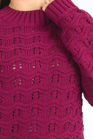 Close-up of someone wearing the Foxwood Carter Knit sweater in magenta, featuring a textured square pattern and ribbed cuffs and neckline. Made from a cotton acrylic blend for softness, with the wearer’s hand visible near the collar.