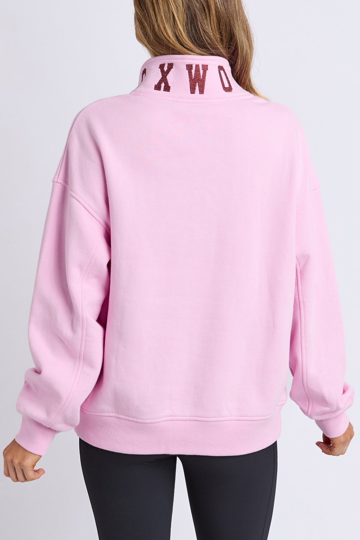 A person wears the Foxwood College Contrast Zip Crew—a light pink 100% cotton relaxed fit jumper with a high collar and dark red "OXWO" letters on the back—paired with black pants, facing away from the camera.