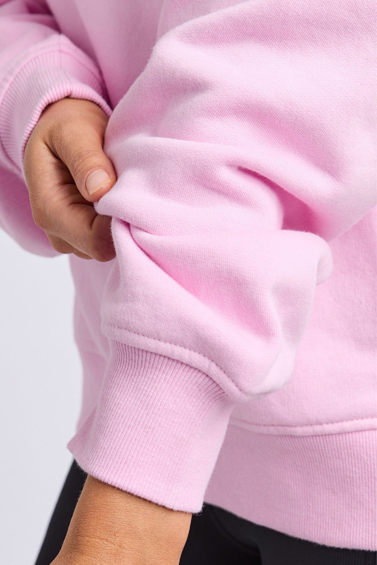 A person is shown from the shoulders down, pulling up a sleeve while wearing the Foxwood College Contrast Zip Crew, a light pink, relaxed fit jumper made from 100% cotton. The background is plain and neutral.