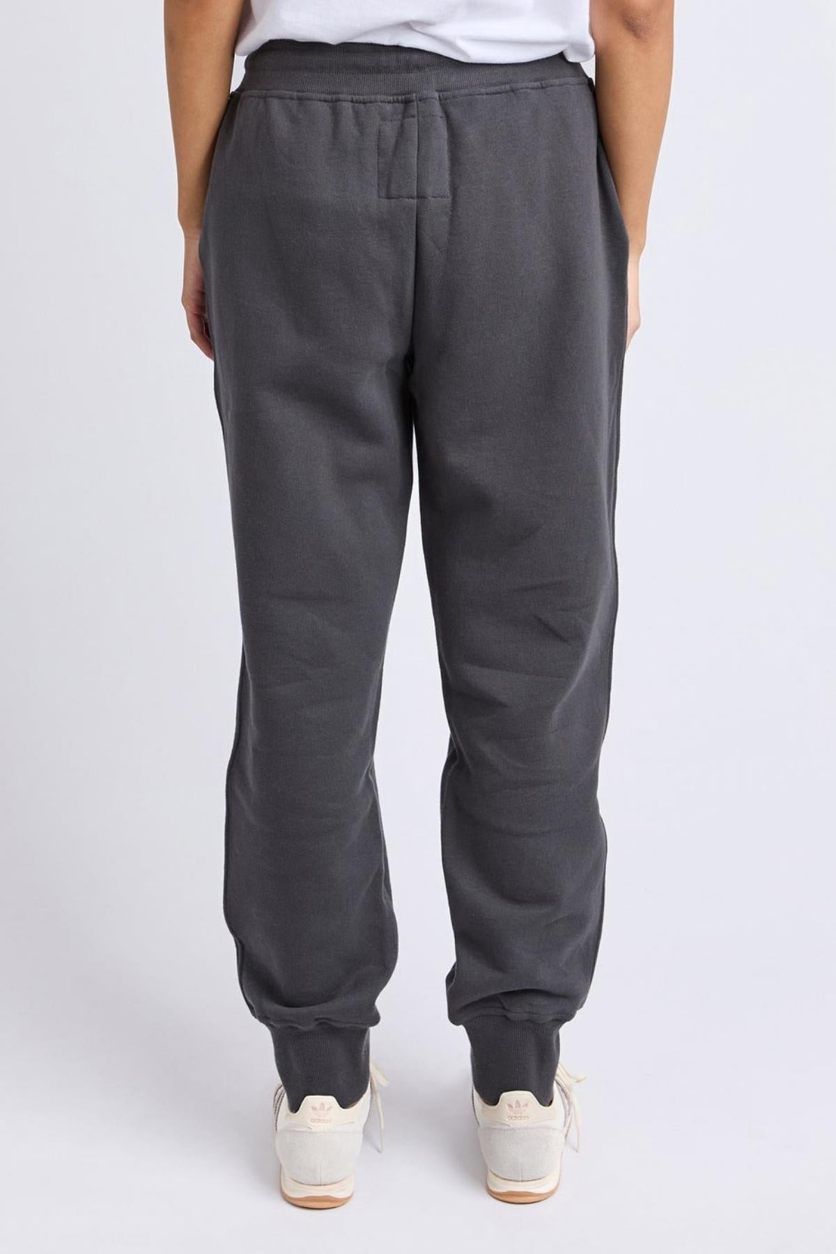 A person stands facing away from the camera, wearing Foxwood College Track Pant in dark gray with an elastic waist, paired with a white shirt and white sneakers, set against a plain light background.
