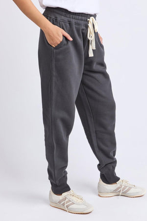Person wearing the Foxwood College Track Pant in dark gray with elastic waist, hands in pockets, white T-shirt partly visible, and cream sneakers, standing against a light background. A timeless silhouette for effortless style.