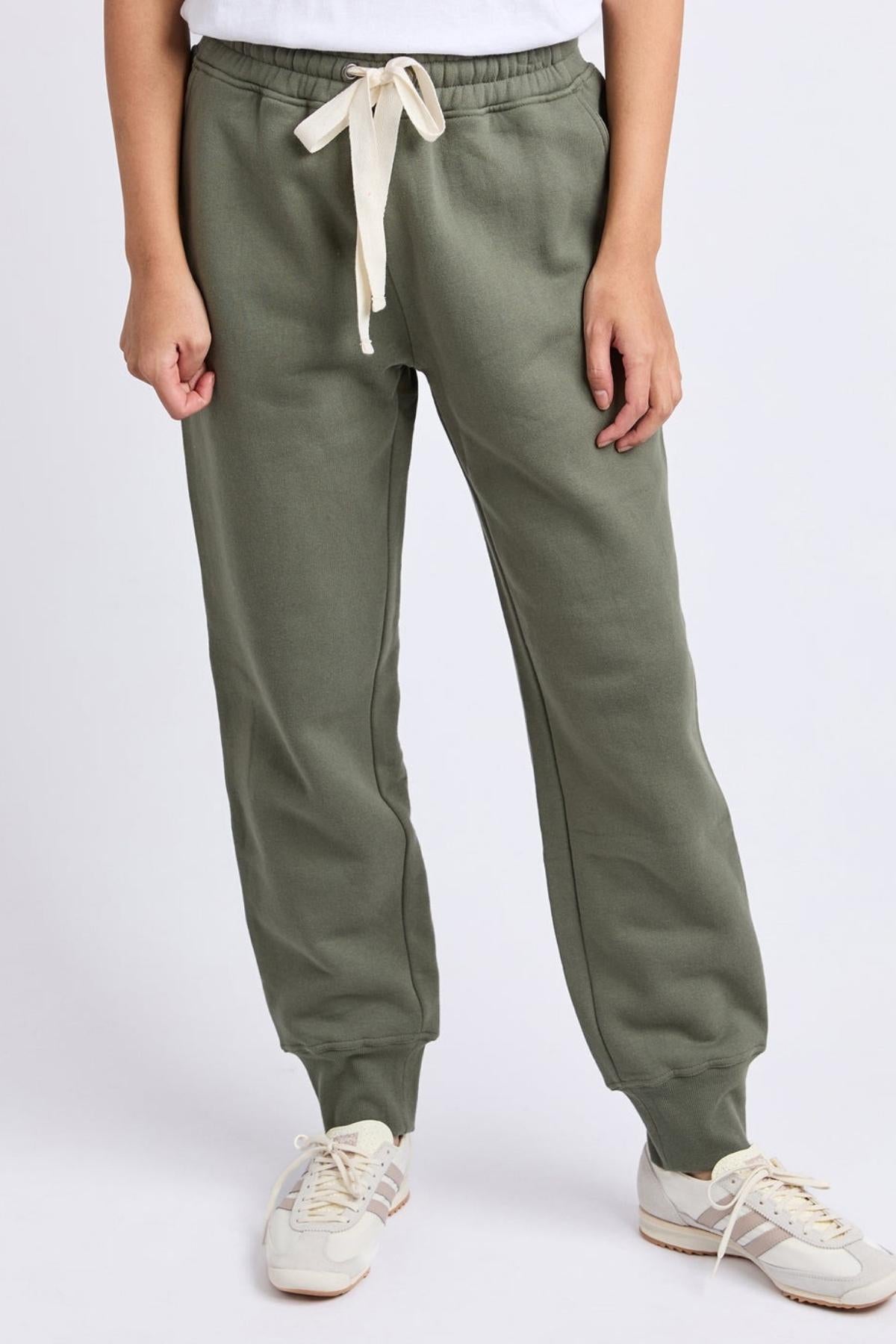 Wearing the Foxwood College Track Pant in olive green with a white drawstring, paired with white sneakers and a white top, someone stands against a plain light background, highlighting both comfort and style.