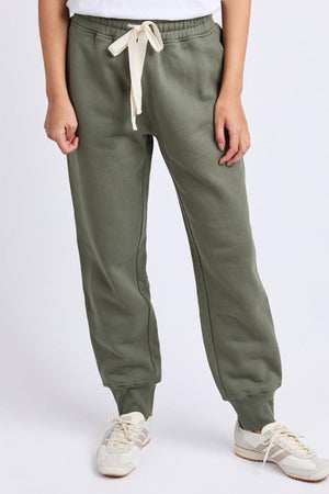 Wearing the Foxwood College Track Pant in olive green with a white drawstring, paired with white sneakers and a white top, someone stands against a plain light background, highlighting both comfort and style.