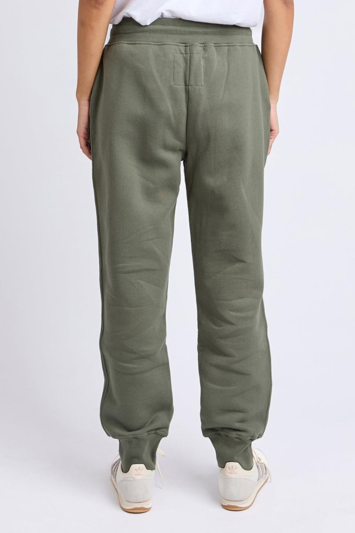 A person faces away, wearing Foxwood College Track Pants in olive green with white sneakers and relaxed hands. The plain light background highlights the pants’ comfort and modern style.
