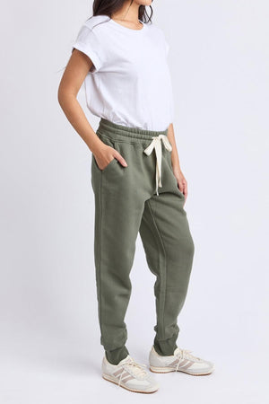 A woman stands with hands in pockets, exuding comfort and style in Foxwood College Track Pant joggers, a white T-shirt, and white sneakers against a plain light background.