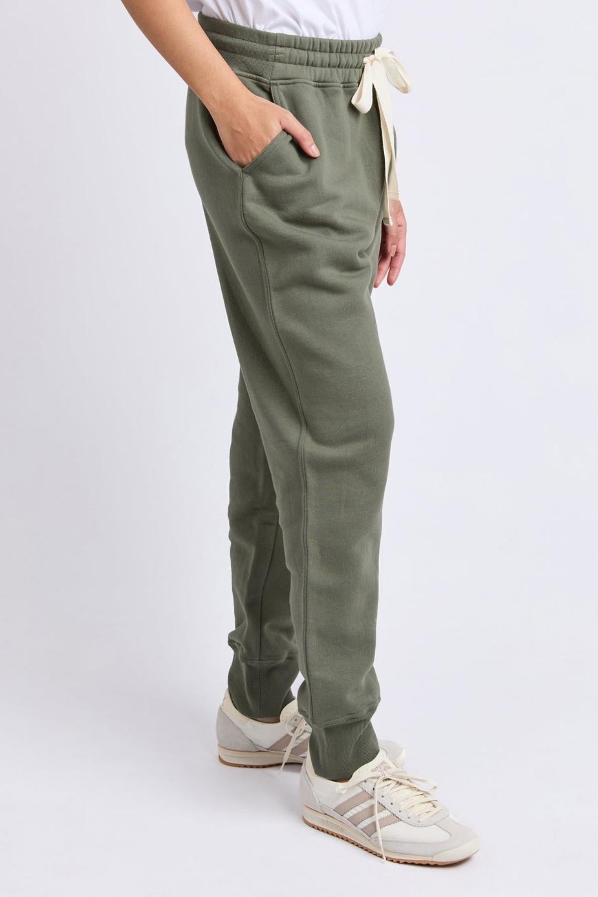 Wearing the Foxwood College Track Pant in olive green for ultimate comfort and style, paired with a white top and white sneakers with beige accents; model stands sideways, one hand in pocket, against a plain background.