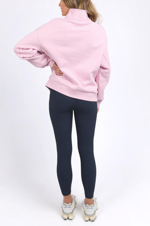 A person faces away from the camera, wearing a soft cotton Foxwood College Zip Crew with an embroidered collar, paired with dark blue leggings and white sneakers, set against a plain white background.