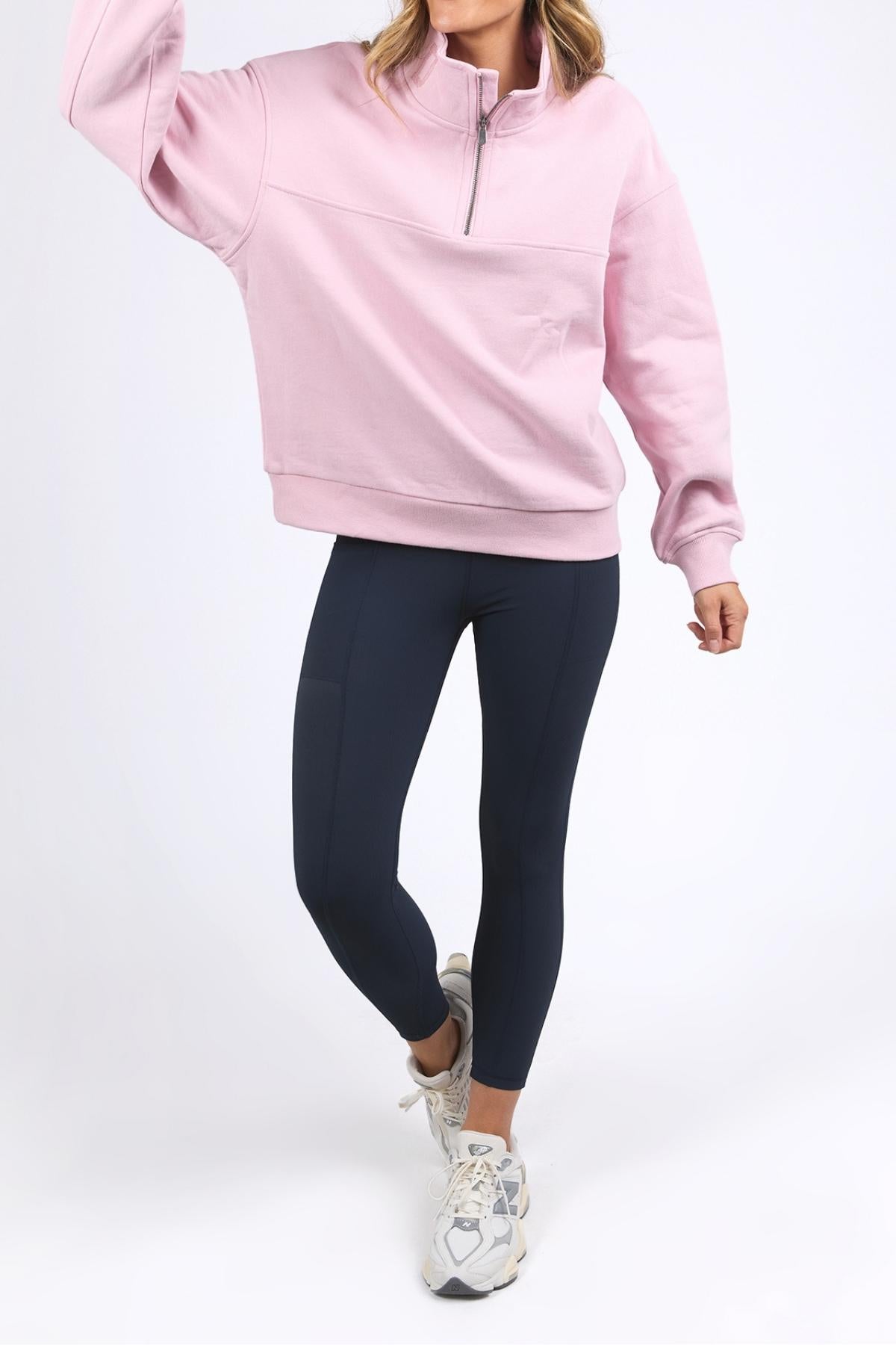 Wearing the light pink Foxwood College Zip Crew, crafted from soft cotton with an embroidered collar, this person pairs it with dark navy leggings and white sneakers while standing against a plain white background. Their face is not shown.
