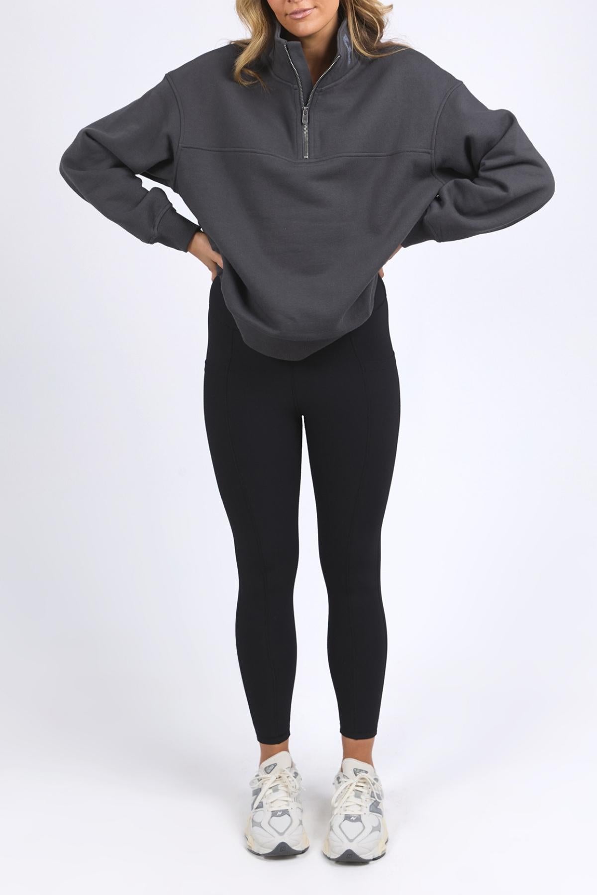 A woman with hands on hips wears a dark gray Foxwood College Zip Crew, black leggings, and white sneakers against a plain white background. Her face is partially out of frame.