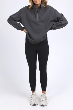 A woman with hands on hips wears a dark gray Foxwood College Zip Crew, black leggings, and white sneakers against a plain white background. Her face is partially out of frame.