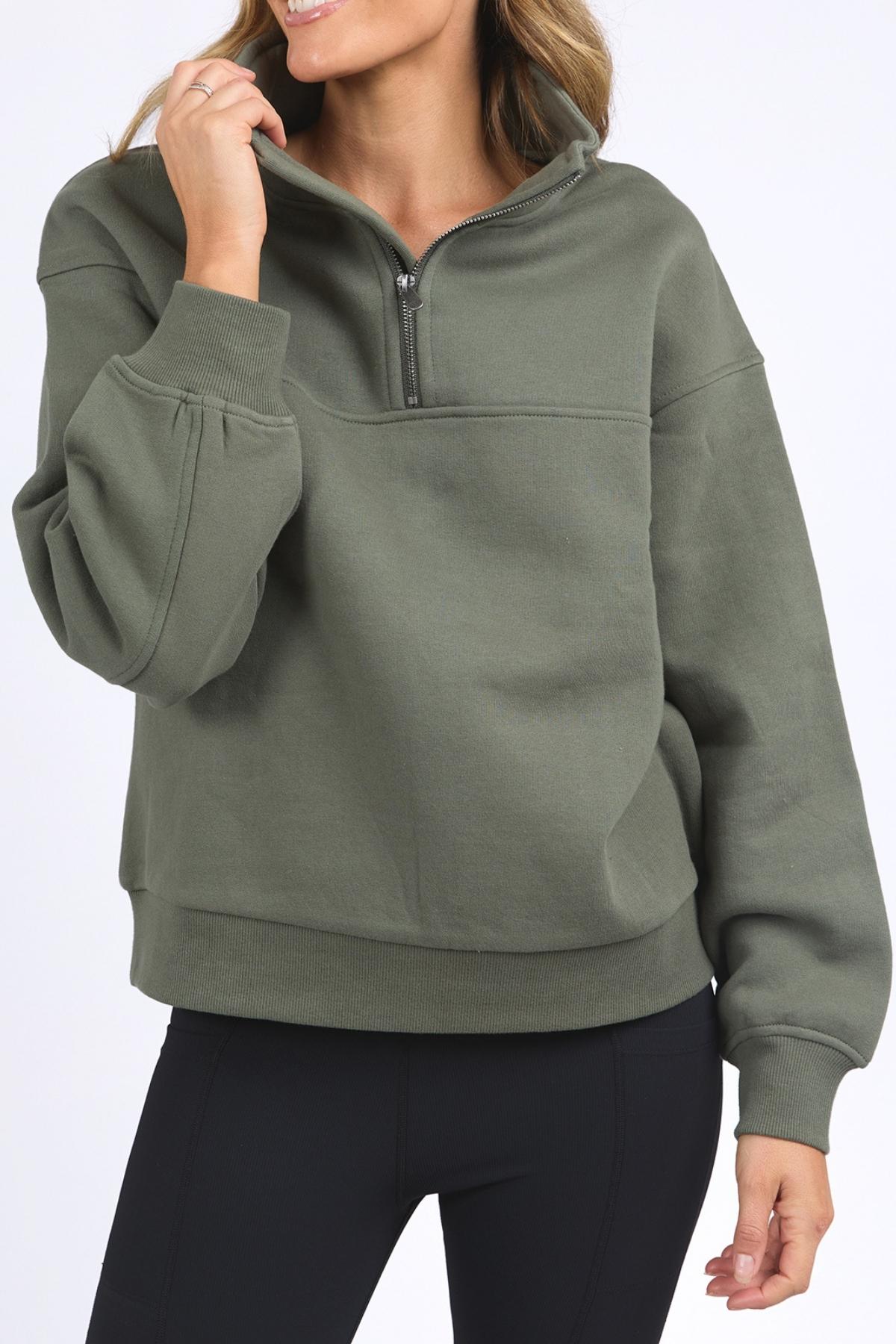 A woman in an olive green Foxwood College Zip Crew and black leggings smiles, holding the pullover's soft cotton collar. The image is cropped at her mouth and mid-thighs.