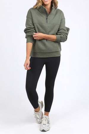 A woman wearing the Foxwood College Zip Crew in green, paired with black leggings and white sneakers, stands against a plain white background, smiling confidently with one hand on her waist.