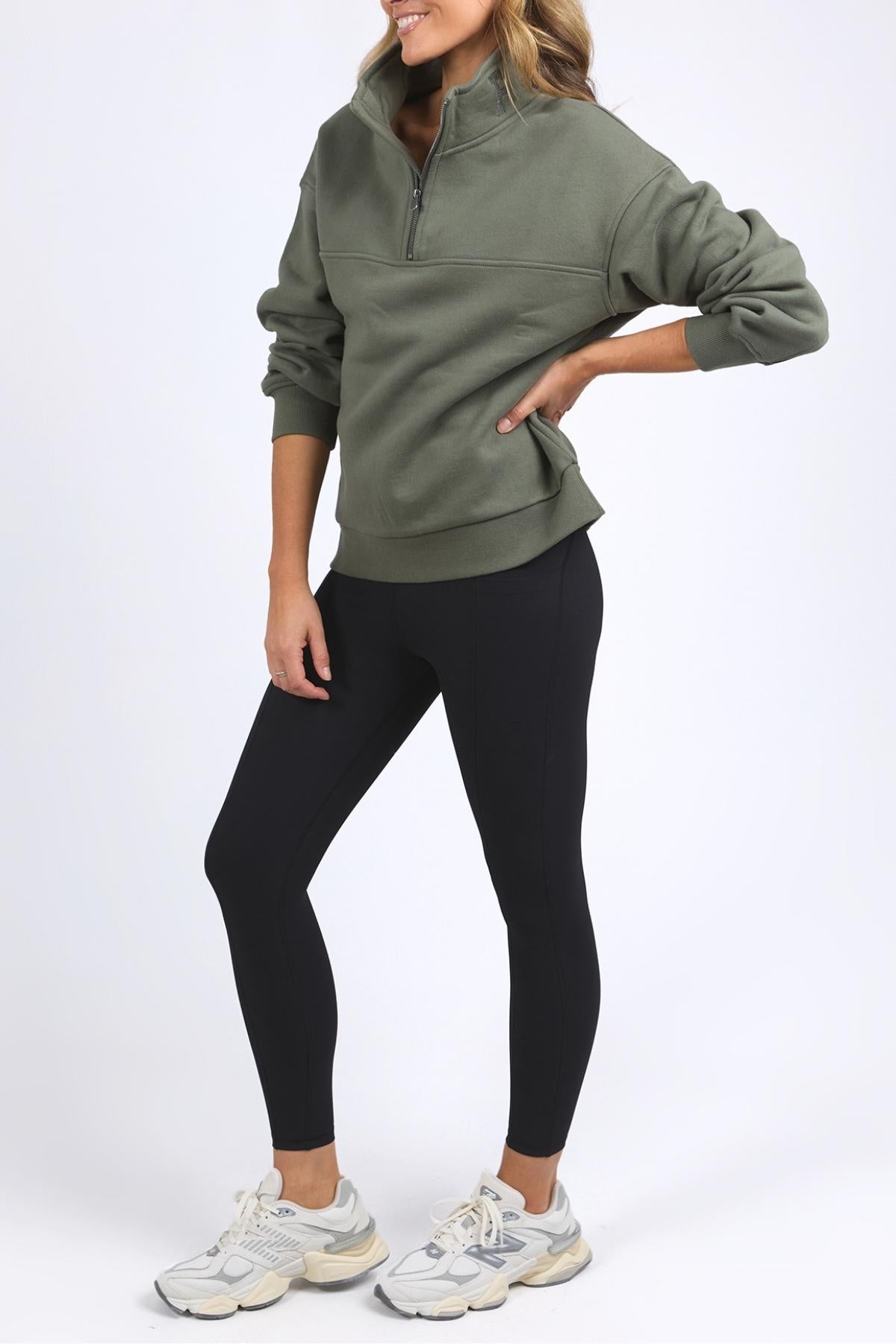 Someone wears a Foxwood College Zip Crew, black leggings, and white sneakers, posing with one hand on their hip in front of a plain white background. Their face is not visible.