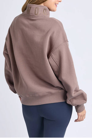 A woman wearing the Foxwood College Zip Crew in soft mauve cotton with "WONDER" on the collar and navy blue leggings is shown from the back, smiling against a plain light background—perfect for casual outings.