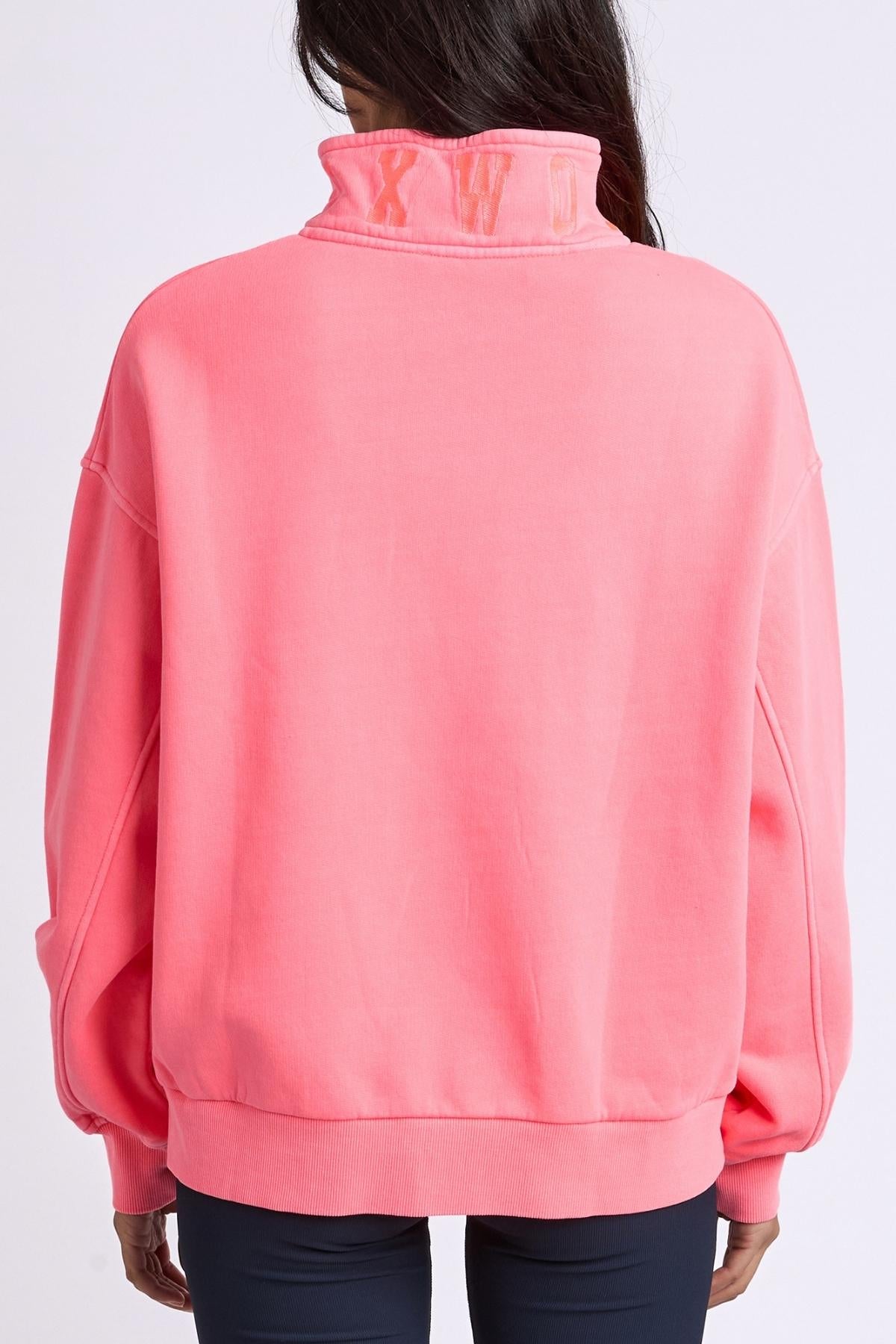 A person with long dark hair faces away from the camera, wearing the Foxwood College Zip Crew in bright pink cotton with a loose fit, high embroidered collar, and subtle lettering, set against a plain background.