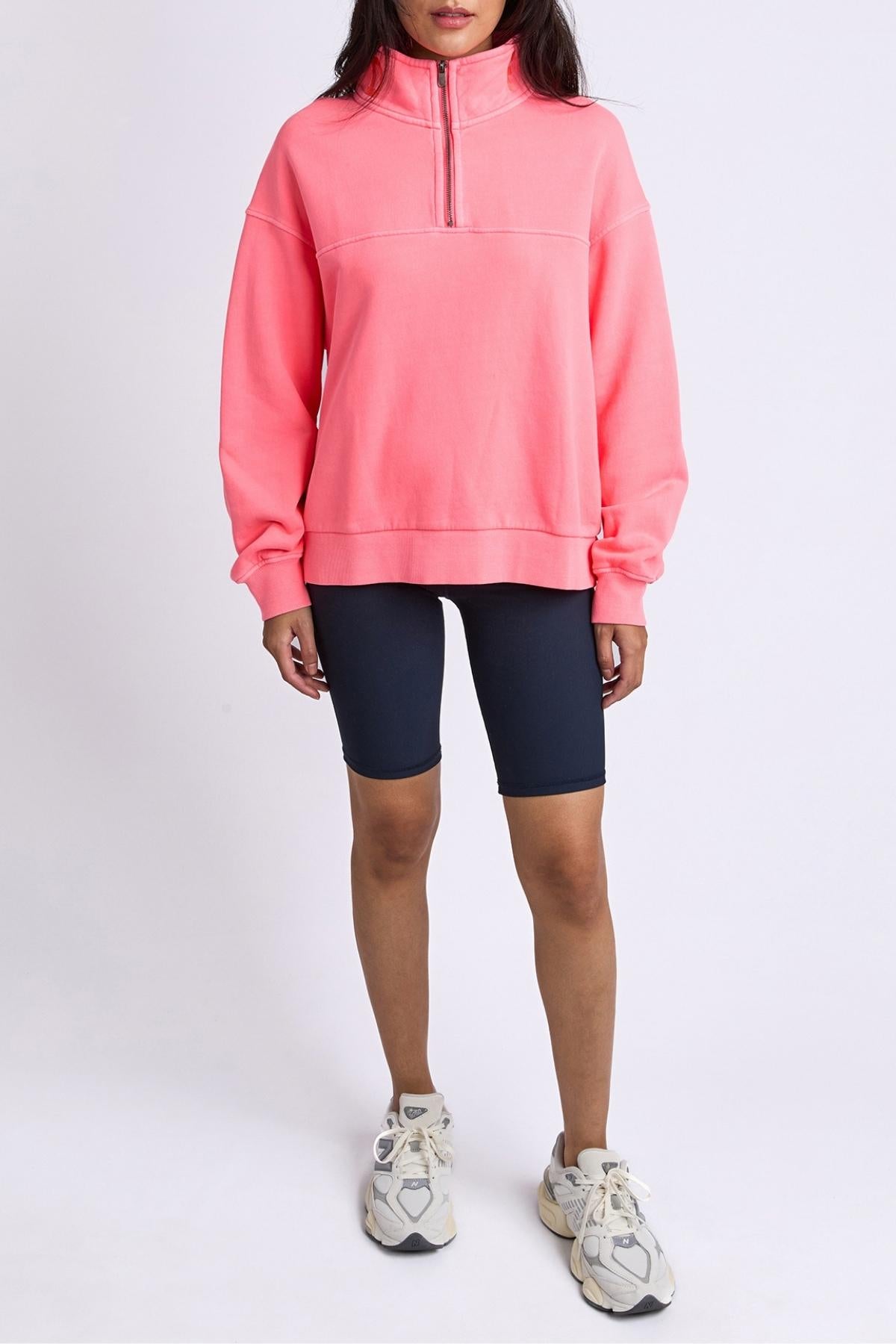 Someone wears the Foxwood College Zip Crew in bright pink, featuring an embroidered collar, paired with dark bike shorts and white sneakers, standing against a plain white background.