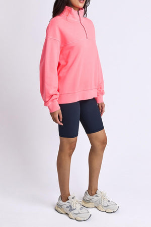 A woman wears the Foxwood College Zip Crew in bright pink, paired with navy biker shorts and white sneakers, standing against a plain white background with her hair down and hands by her sides.