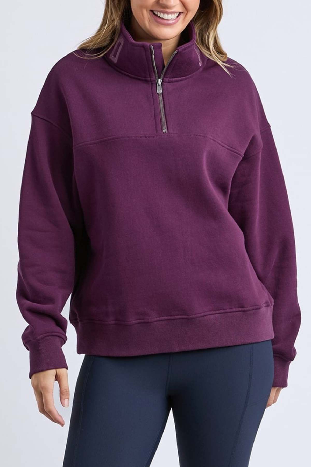 A person smiles with hands by their sides, wearing dark leggings and the Foxwood College Zip Crew, a purple quarter-zip pullover sweatshirt with an embroidered collar, standing against a plain light background.