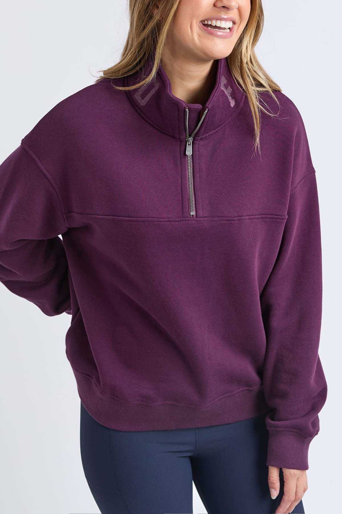 A woman smiles with one hand on her hip, wearing the Foxwood College Zip Crew—a purple pullover with an embroidered collar, high quarter-zip, and made from 100% soft cotton—set against a plain light background.