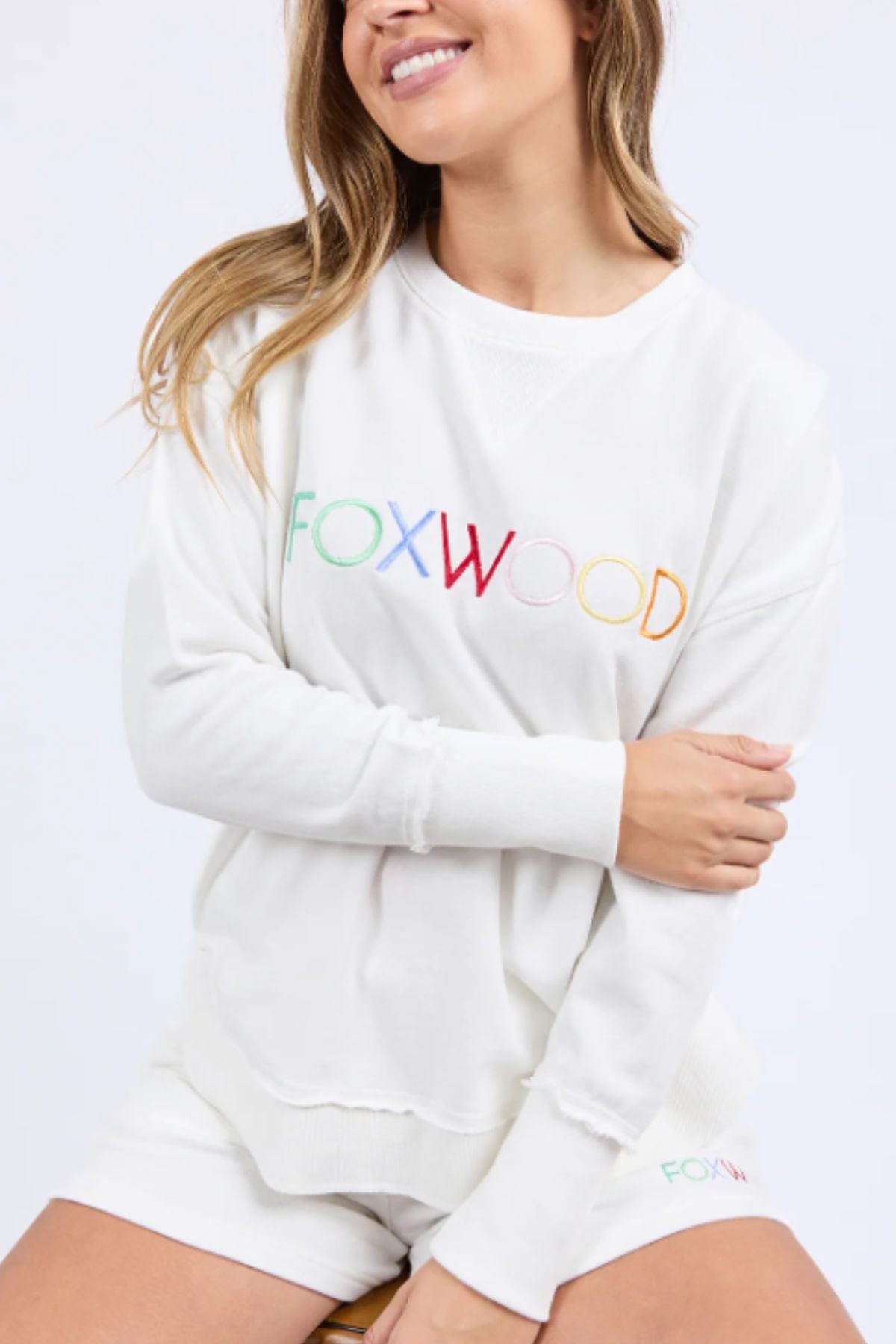 A woman sits on a stool, smiling with her arms loosely crossed, wearing the Foxwood Simplified Confetti Crew—a white 100% cotton sweatshirt and shorts set with "FOXWOOD" in multicoloured embroidery across the front.
